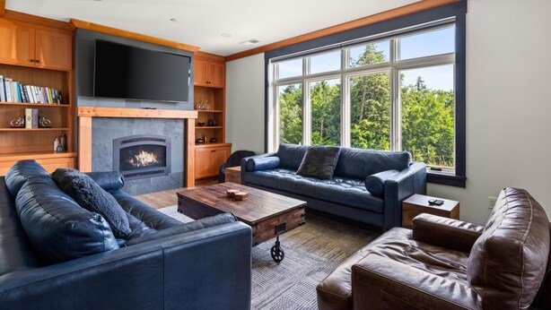 A modern living room with a fireplace, leather sofas, a coffee table, and a large window with a view of trees. - Stunning Trapp Lodge Townhouse, Brand New in Stowe (Stowe)