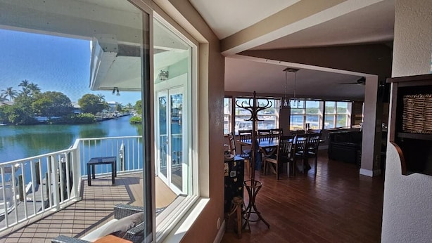 A spacious room with a large sliding glass door leading to a balcony overlooking a body of water. Inside, there is a dining area with wooden flooring and a table set for two.