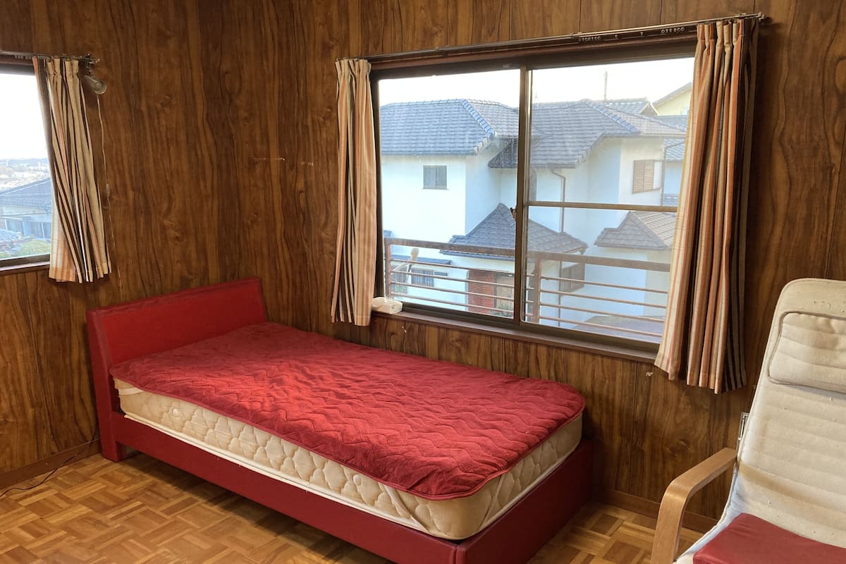 A room with a single bed, wooden walls, a window with curtains, and a chair.