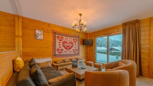 Ein gemütliches Blockhaus-Interieur mit einer Couch, Sesseln, einem Couchtisch, einer Kristallleuchtre und einem Fernseher, sowie einem großen Fenster mit Blick auf schneebedeckte Bäume.