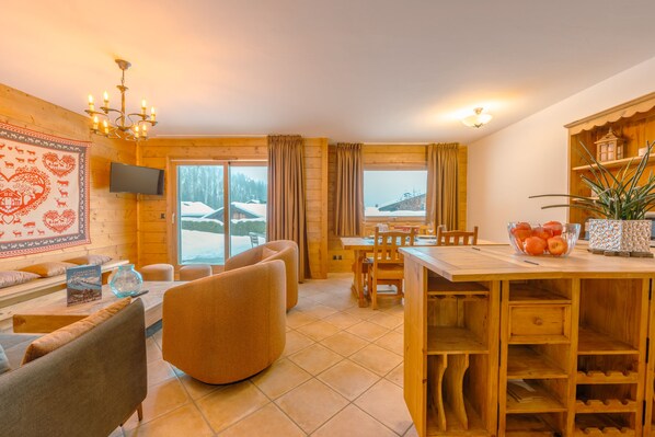 Une pièce chaleureuse avec un décor intérieur en bois, une table à manger, des chaises, un canapé, un téléviseur et une fenêtre donnant sur un paysage enneigé.