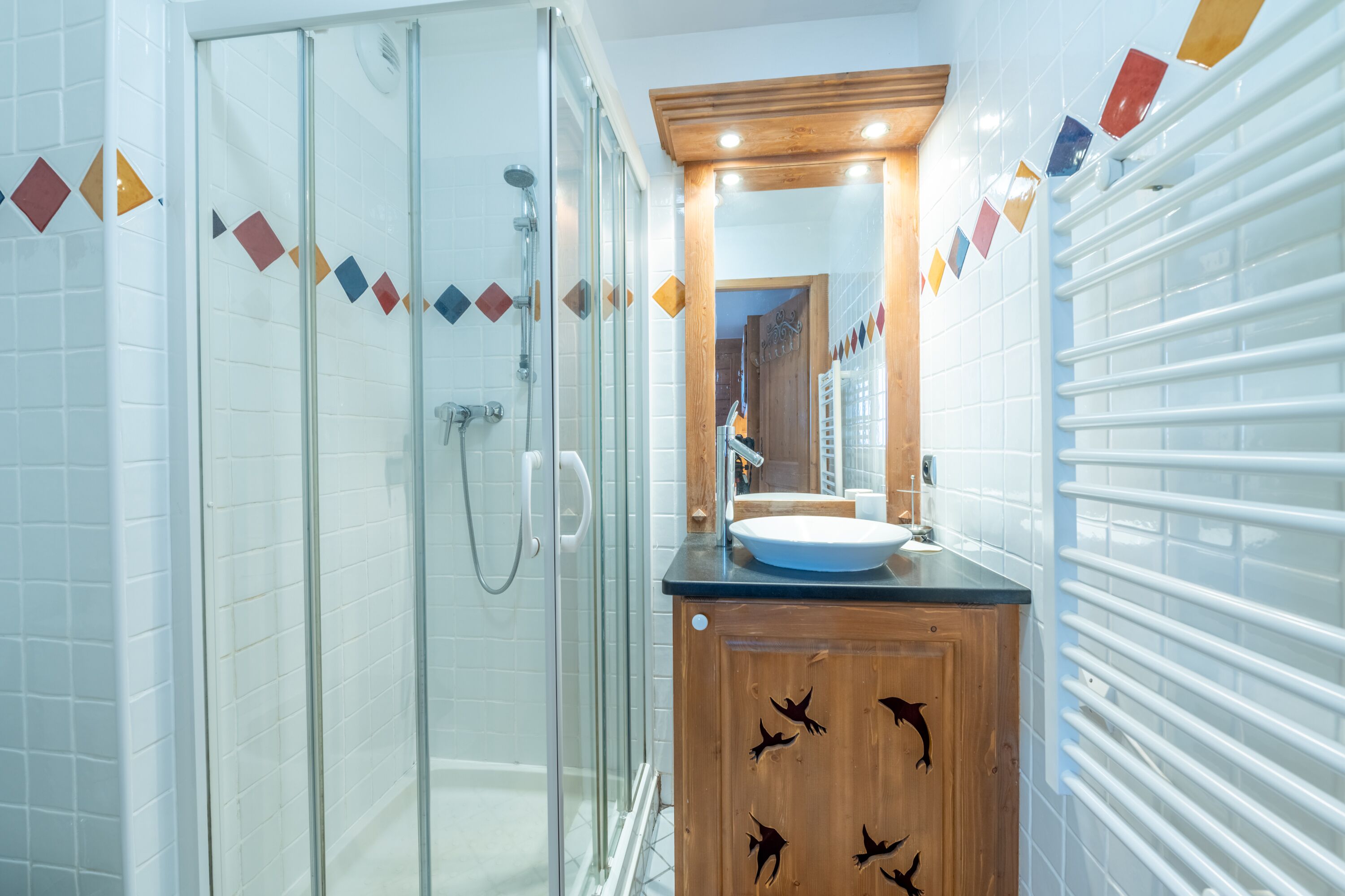 Une salle de bain moderne dotée d’une douche à parois de verre, d’un meuble-lavabo en bois et d’un mur carrelé orné de motifs géométriques en forme de losanges.