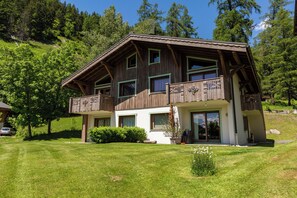Une maison en bois à deux étages, dotée d’un balcon, entourée d’une pelouse bien entretenue et d’arbres.