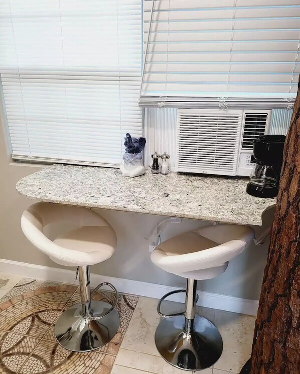 A kitchen with a granite countertop, two white bar stools, a coffee maker, and a window with blinds.
