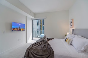Un dormitorio moderno con cama, televisor en la pared y vistas a un paisaje urbano a través de un ventanal grande.
