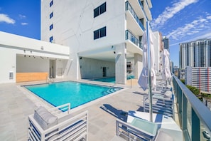 Una piscina en la azotea con vistas despejadas al horizonte urbano, una zona de estar techada y un edificio moderno de líneas geométricas.