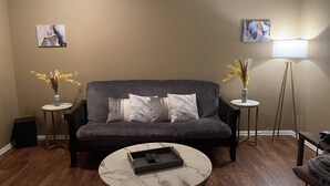 Ein Wohnzimmer mit einem dunkelgrauen Sofa, zwei Beistelltischen mit Vasen, einer Stehlampe und einem Couchtisch mit Fernbedienung.