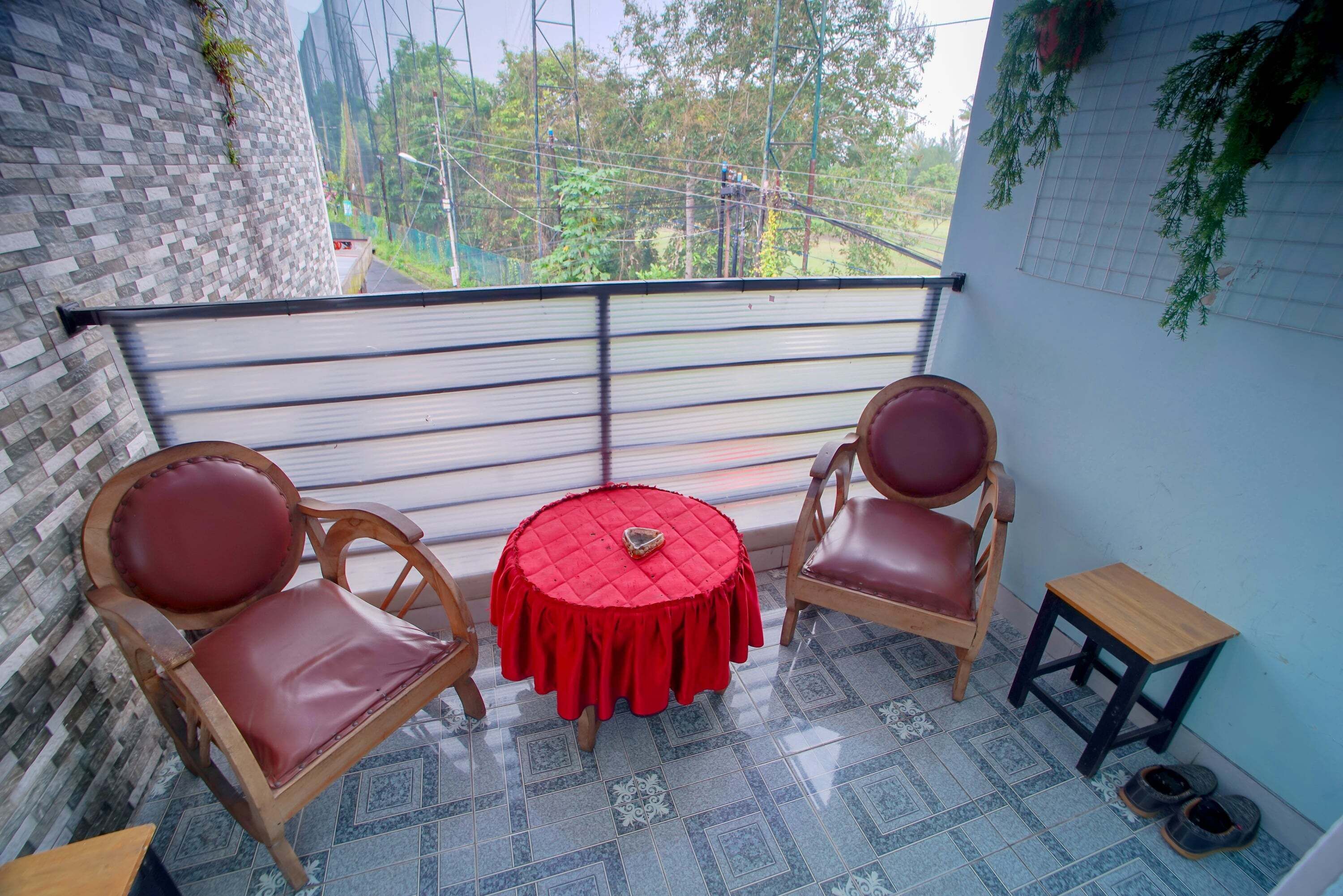 A balcony with two wooden chairs and a small table, a tiled floor, and a view of greenery and buildings in the background.