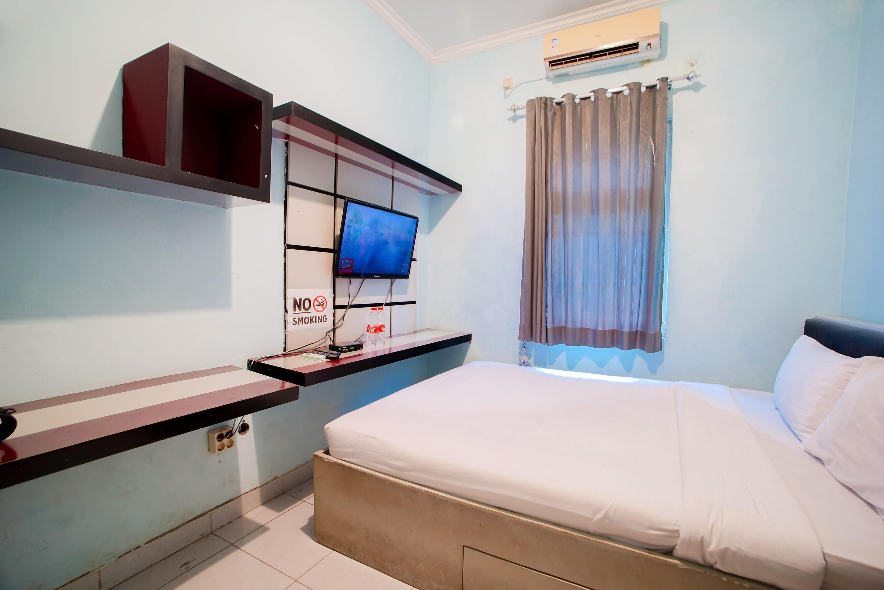 A hotel room with a bed, a TV mounted on the wall, a no-smoking sign, and an air conditioning unit.