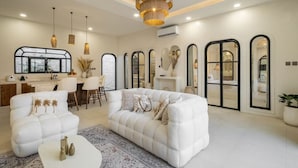 Un salón amplio con un sofá blanco, una mesa de centro y un comedor con sillas y una mesa. Hay ventanas grandes y una araña.