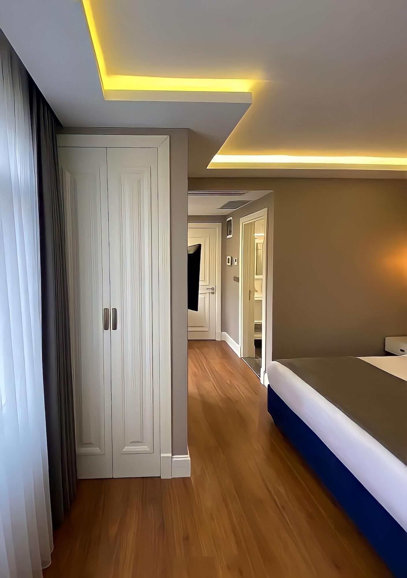 Quarto de hotel moderno com piso de madeira, uma cama, um guarda-roupa e um corredor que leva a outro quarto.