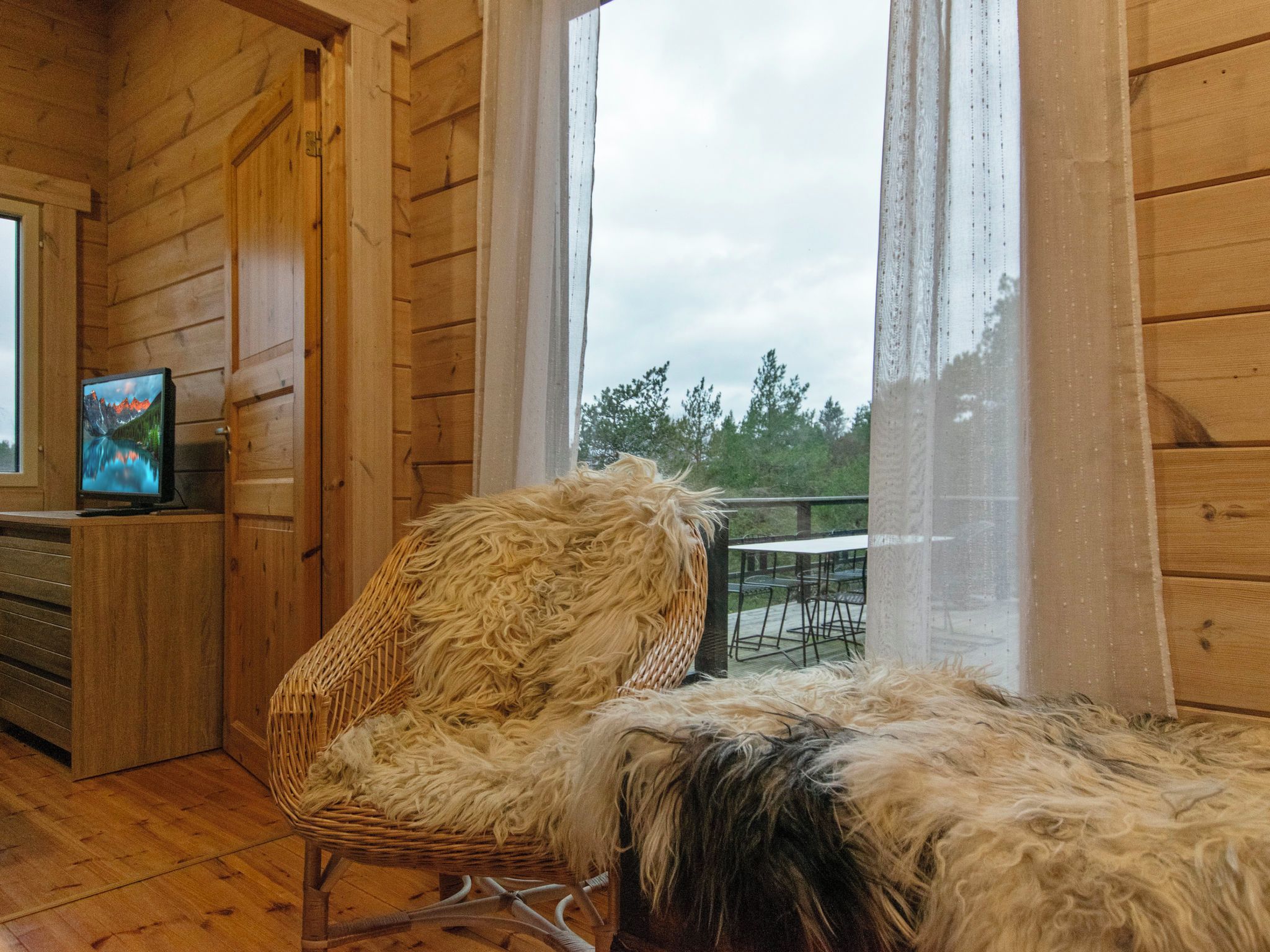 A wooden cabin interior with a wicker chair, a television on a shelf, and a view of trees through a window.