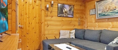 Een knus interieur met een houten wand, een bank, een glazen bijzettafeltje en een schilderij van een schip.