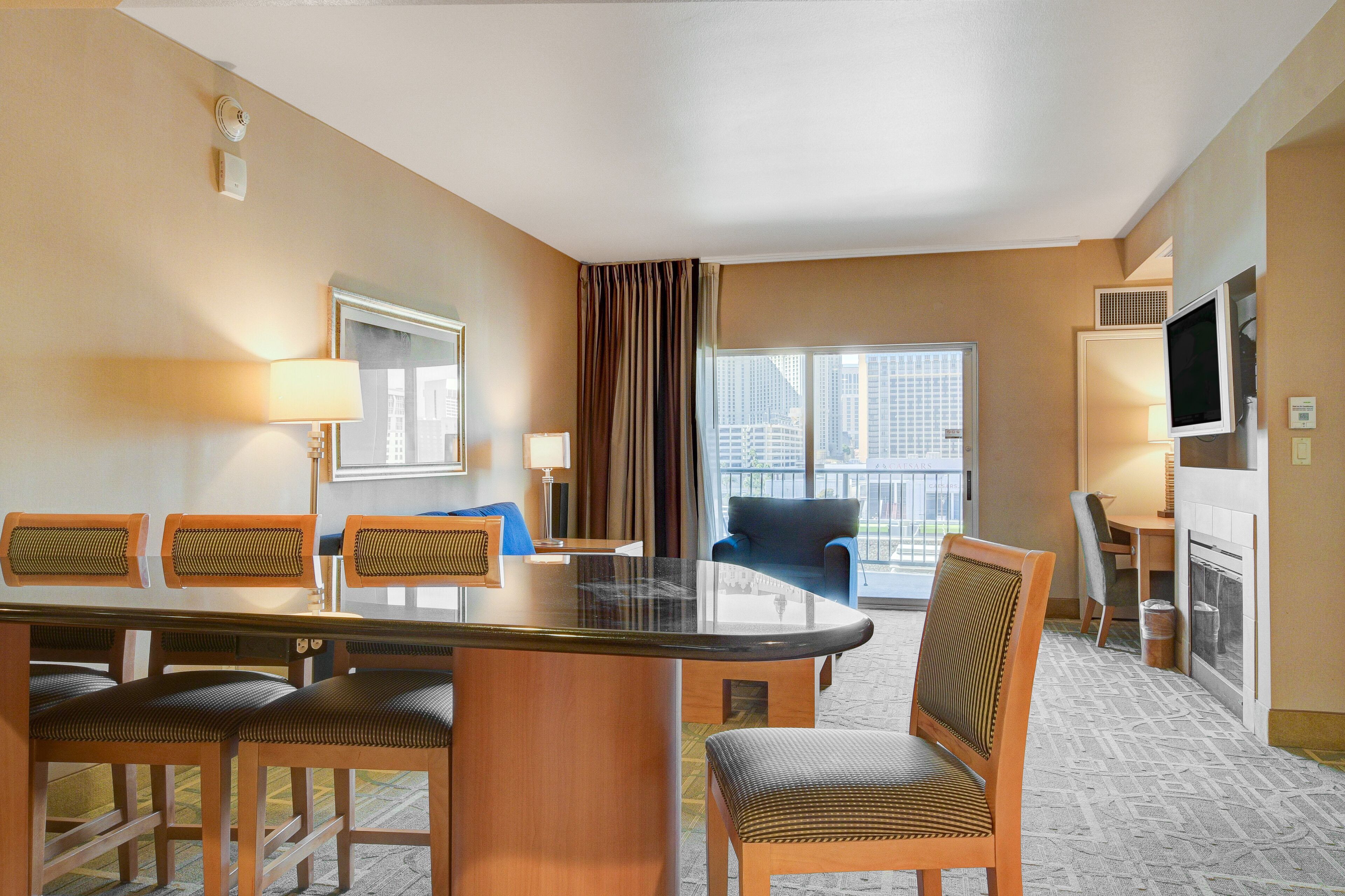 A hotel room with a dining area featuring a glass-top table, wooden chairs, and a view of the city through large windows.
