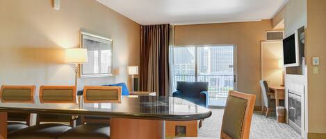 A hotel room with a dining area featuring a glass-top table, wooden chairs, and a view of the city through large windows.