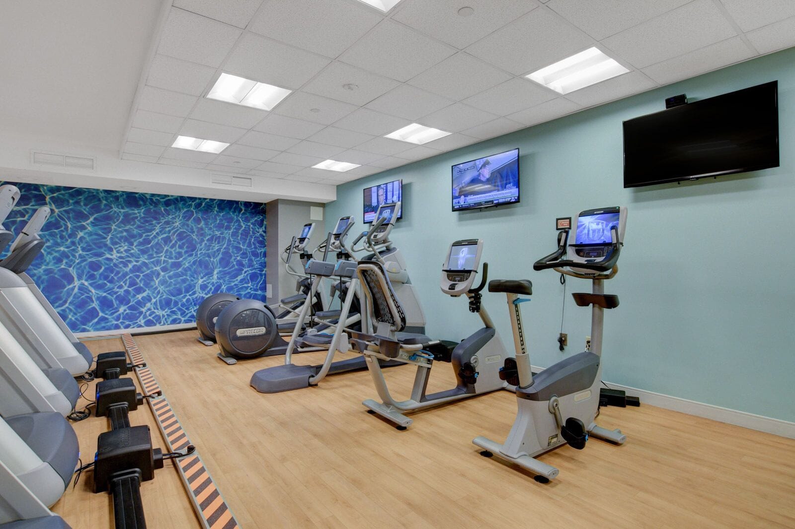 Ein Fitnessstudio mit verschiedenen Cardiogeräten, einem Pool und einem Fernseher, der ein Trainingsprogramm zeigt.