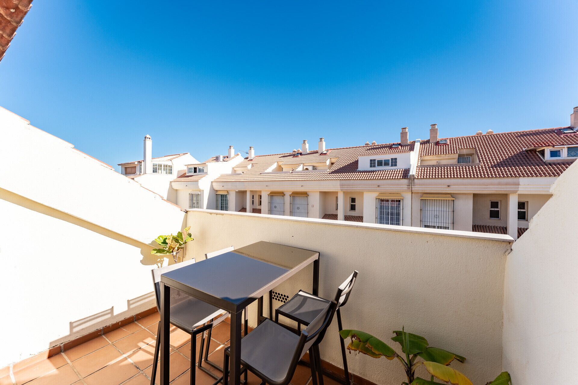 Une terrasse avec une table et des chaises, d’où l’on peut admirer une rangée de bâtiments blancs aux toits recouverts de tuiles rouges.