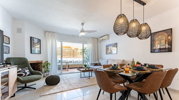 Ein modernes Wohnzimmer mit Essbereich, ausgestattet mit einer Couch, einem Couchtisch und einem Esstisch für vier Personen. Es gibt einen Deckenventilator, einen Kamin und ein großes Fenster mit Blick ins Freie.