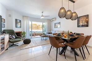 A modern living room with a dining area, featuring a sofa, a coffee table, and a dining table set for four. There is a ceiling fan, a fireplace, and a large window with a view of the outdoors. - Stylish Seaview Townhouse: 3BR, Pool, 4 Terraces REF030 (Benalmádena)