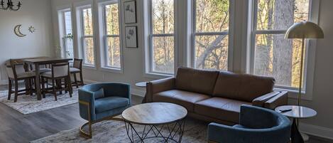 A spacious living room with a brown sofa, blue armchairs, a round coffee table, and a dining area with a table and chairs. Large windows provide natural light and views of trees.