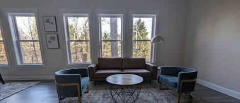 Uma sala de estar ampla com uma grande janela, um sofá bege, duas poltronas e uma mesa de centro.