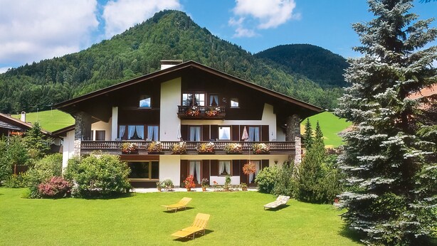 Een tweelaags chalet met een balkon, een verzorgd gazon en een bergachtige achtergrond.