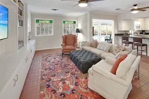 A spacious living room with a large sectional sofa, a tufted ottoman, and a patterned rug. There are two windows with shutters, a ceiling fan, and a kitchen area visible through an open door.