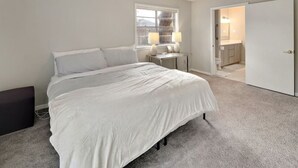 A bedroom with a large bed, a nightstand, a window, and a door leading to a bathroom. - Poolside Paradise - Historic District Home (College Station)