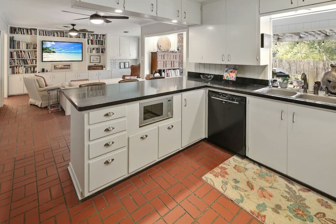A modern kitchen with white cabinetry, a black countertop, and a built-in dishwasher. There is a TV mounted on the wall and a bookshelf filled with books.