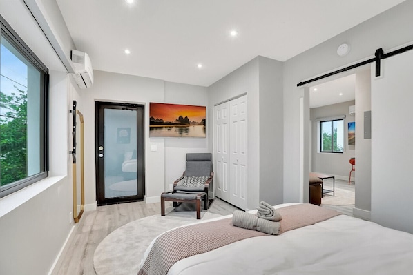 A modern bedroom with a large bed, a chair, a glass door, and a painting of a sunset.