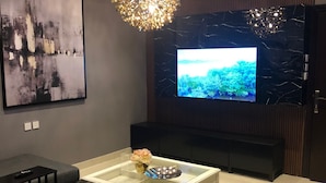 A modern living room with a large flat-screen TV, a glass coffee table, a dark sofa, and a decorative chandelier. - DANA HOTEL RESIDENCES QASSIM (Buraydah)