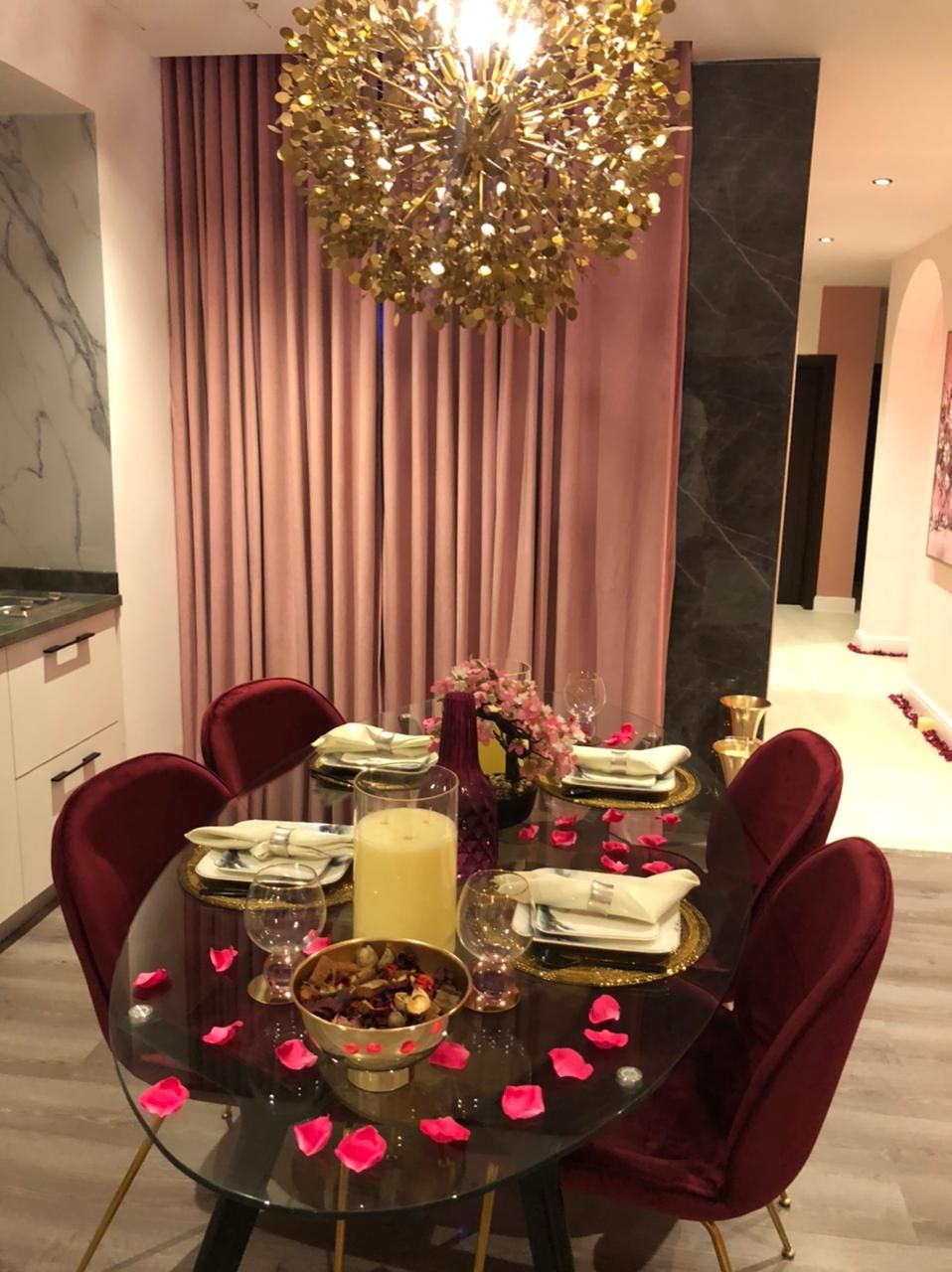 A dining area with a round glass table, red velvet chairs, a gold chandelier, and a backdrop of pink curtains.