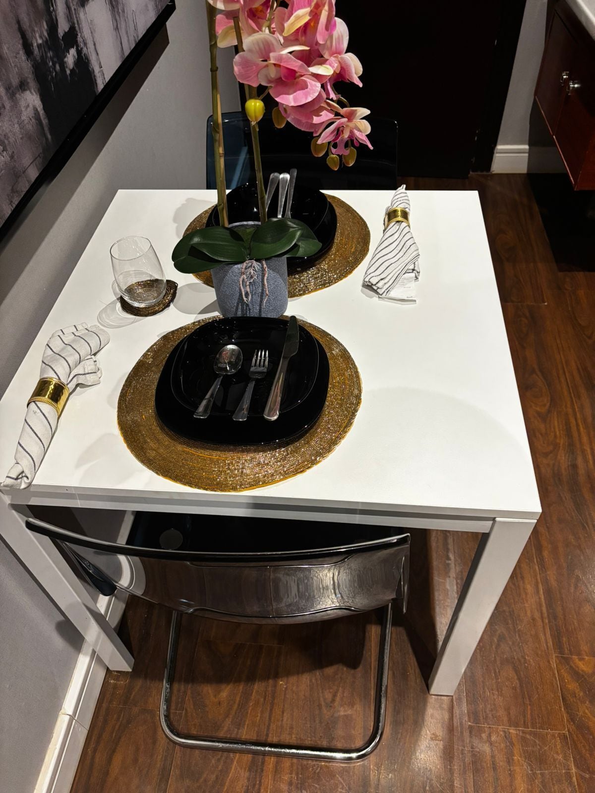 A dining table set with black plates, gold-rimmed placemats, a vase with pink flowers, and a glass of water.