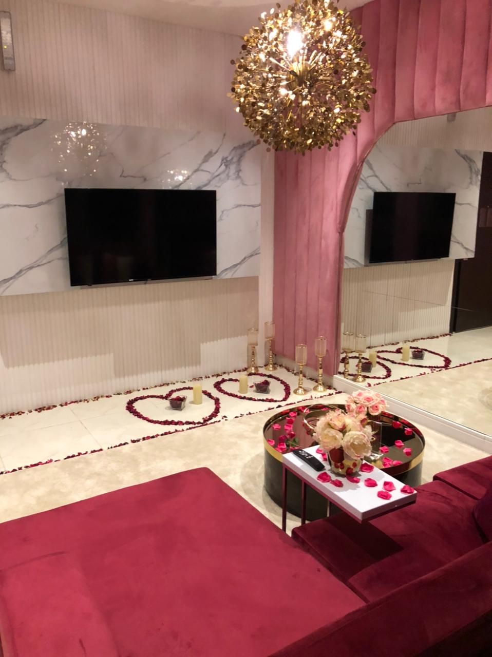 A modern living room with a large flat-screen TV, a decorative chandelier, a heart-shaped flower arrangement, and a red sofa.