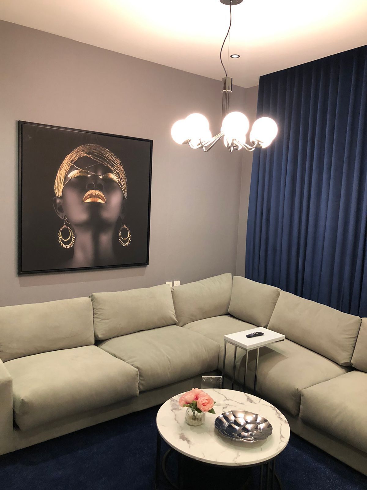 A modern living room with a beige sectional sofa, a round marble table with a vase of flowers, a framed artwork on the wall, and a blue curtain.