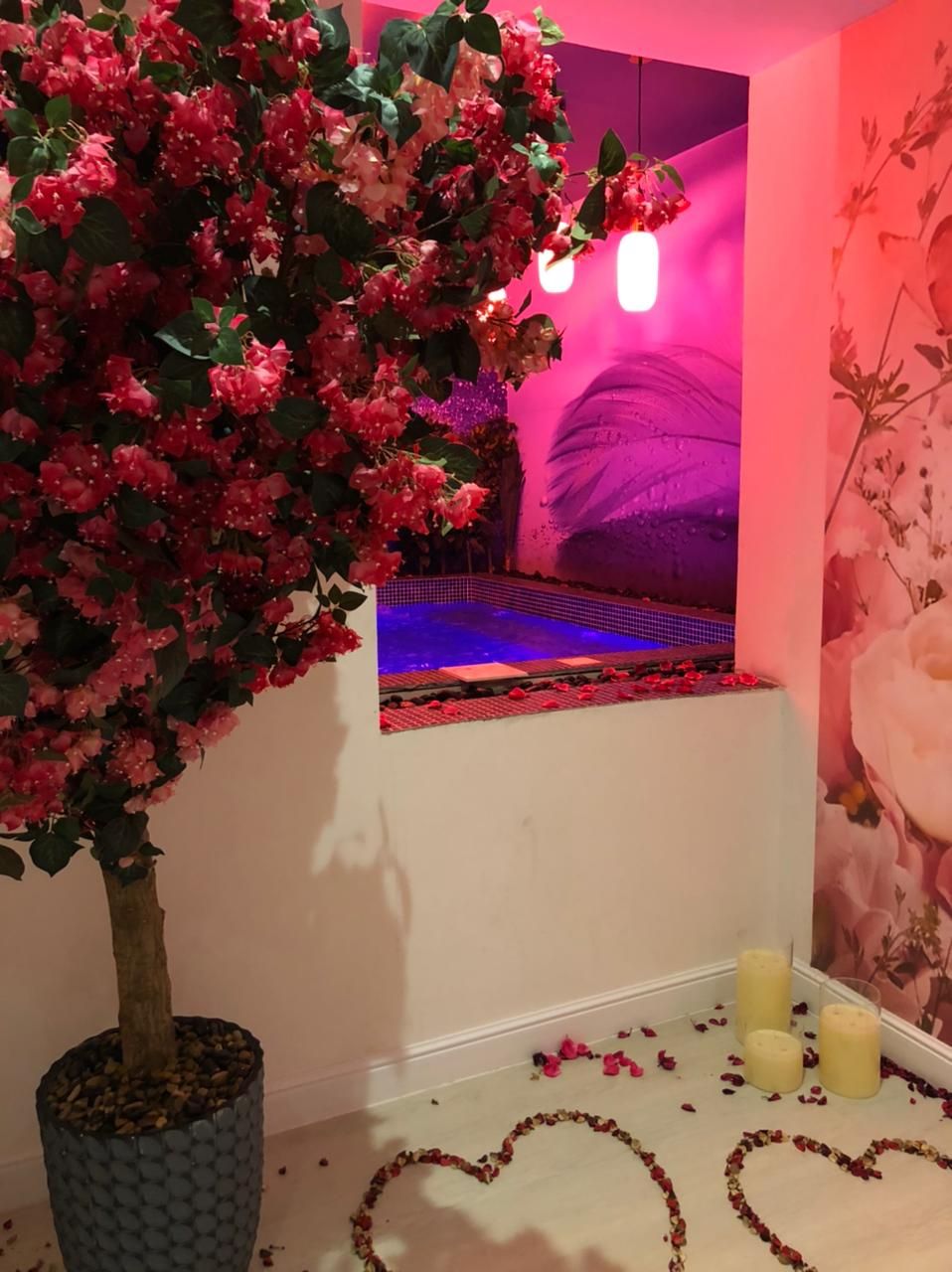 Une pièce avec un grand arbre artificiel, une piscine intégrée au mur et des compositions florales en forme de cœur disposées sur le sol.