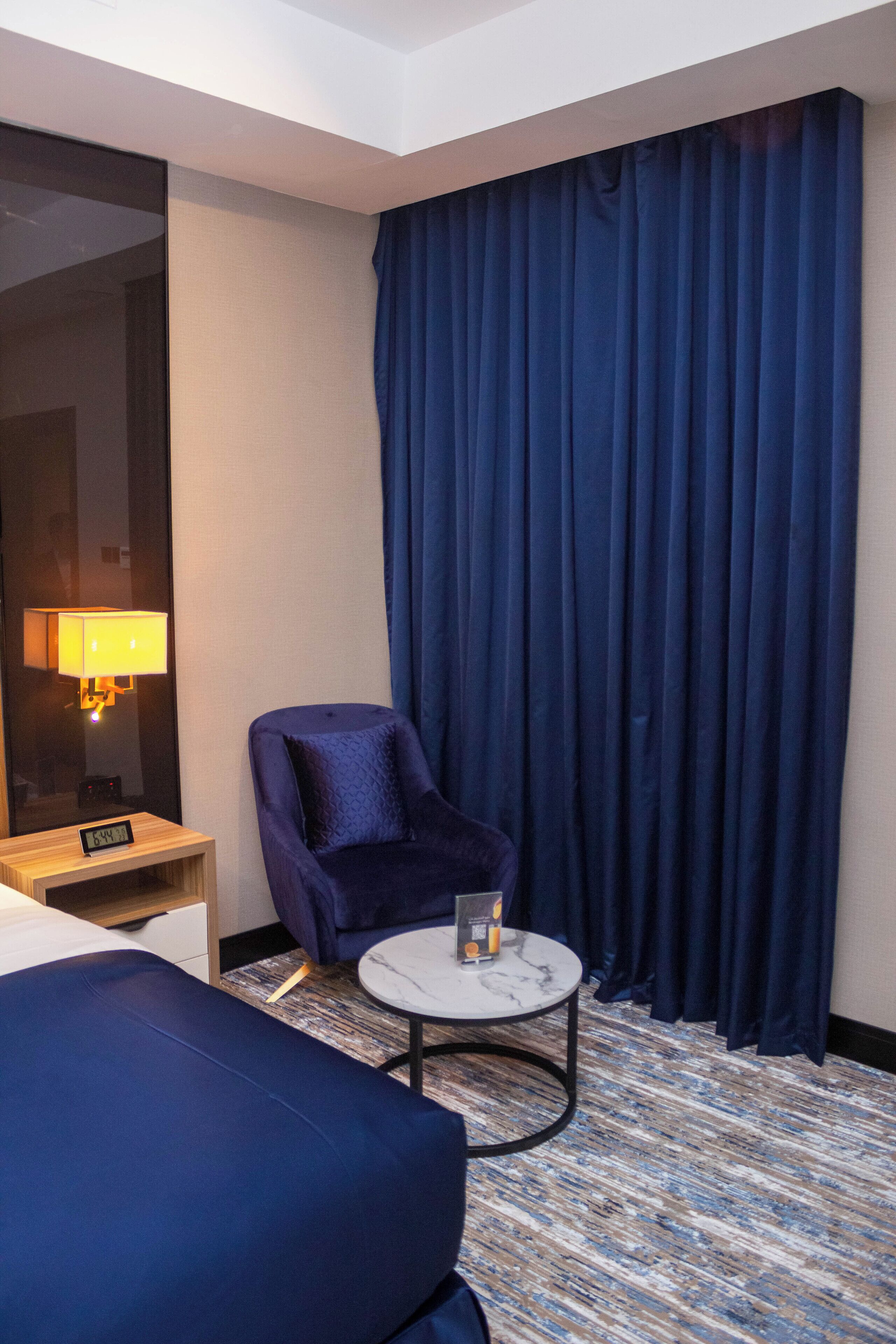 A hotel room with a bed, a chair, a small table, a lamp, and a large window with curtains.