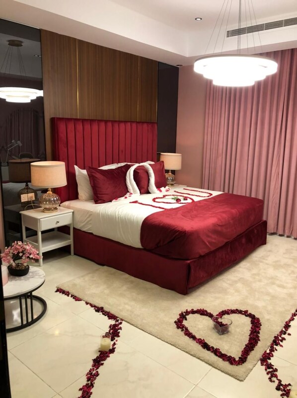 A modern bedroom with a large bed, red bedding, a bedside table with a lamp, and a heart-shaped decoration on the floor. - DANA HOTEL RESIDENCES QASSIM (Buraydah)