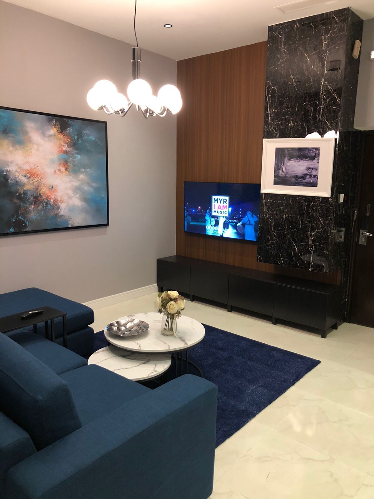 A modern living room with a large TV, blue sofas, a marble coffee table, and a decorative vase.