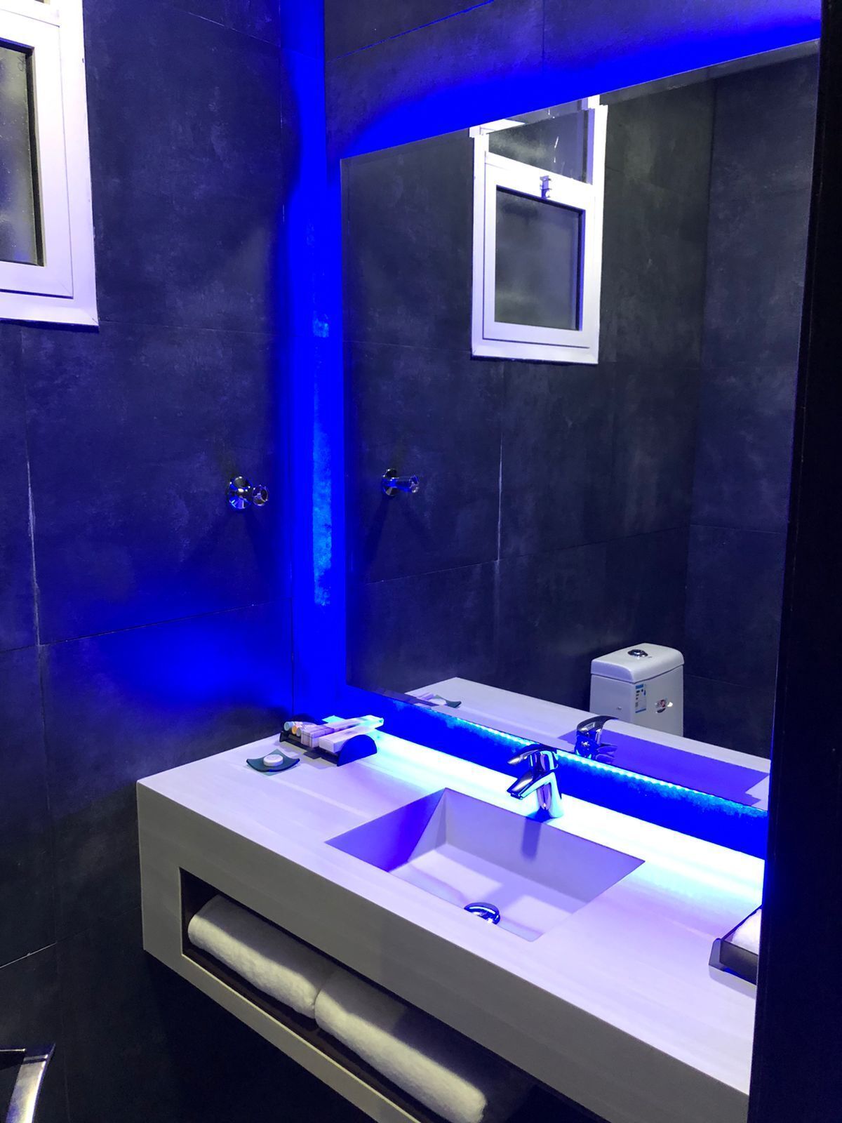 A modern bathroom with a dark tiled wall, a large mirror, and a white vanity with a blue-lit sink.