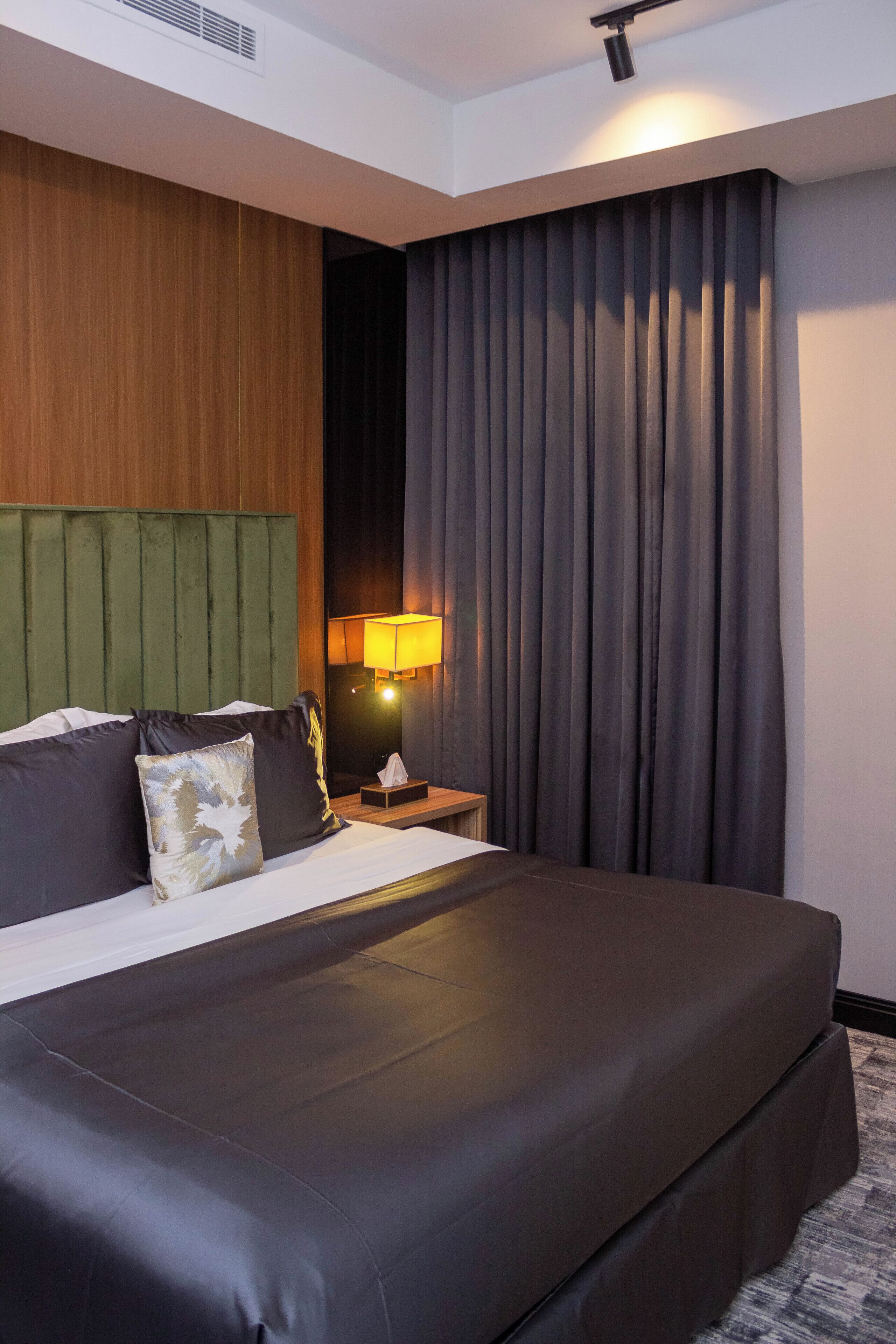 A hotel room with a large bed, a wooden headboard, a nightstand with a lamp, and dark curtains.