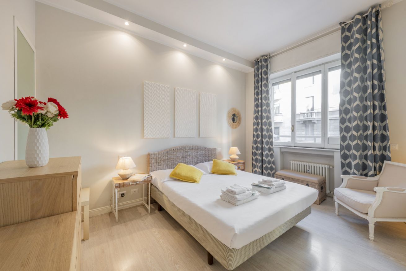 Ein Hotelzimmer mit Bett, Nachttischen, einem Sessel und einem Fenster mit Vorhängen sowie Blick nach außen.