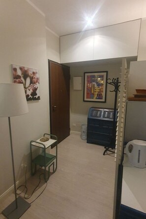 A hallway with a door, a small table with a lamp, a vending machine, and a trash bin. - BnButler - Londonio, 25 - Saddleback (Milano)
