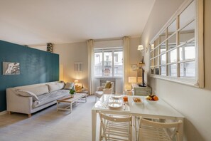 Una sala de estar moderna con sofá, mesa de comedor y sillas, vista de la ciudad a través de amplios ventanales y una pared de acento color turquesa.