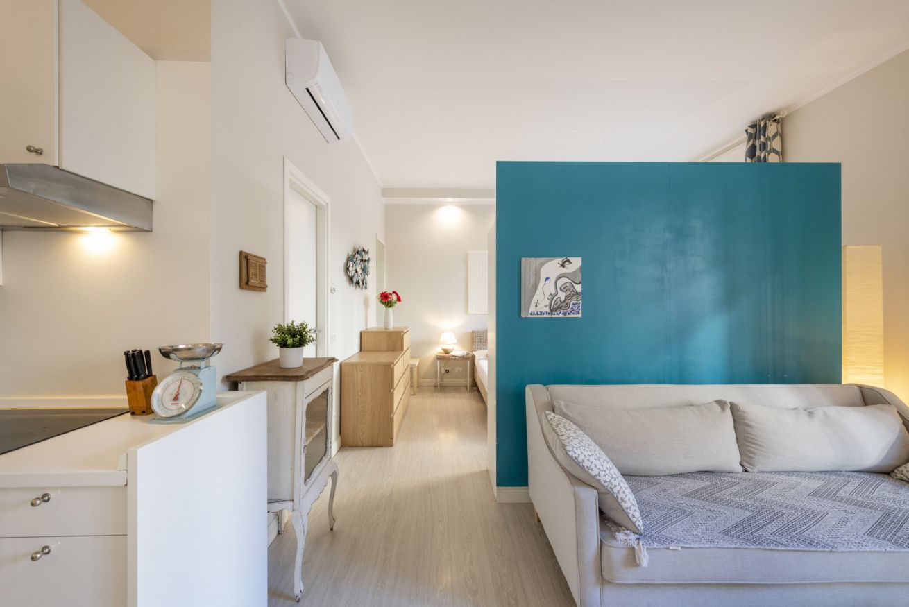 Une cuisine moderne avec des meubles blancs, une balance et une petite plante. Le salon est agrémenté d’un mur d’accent de couleur turquoise et d’un canapé blanc.