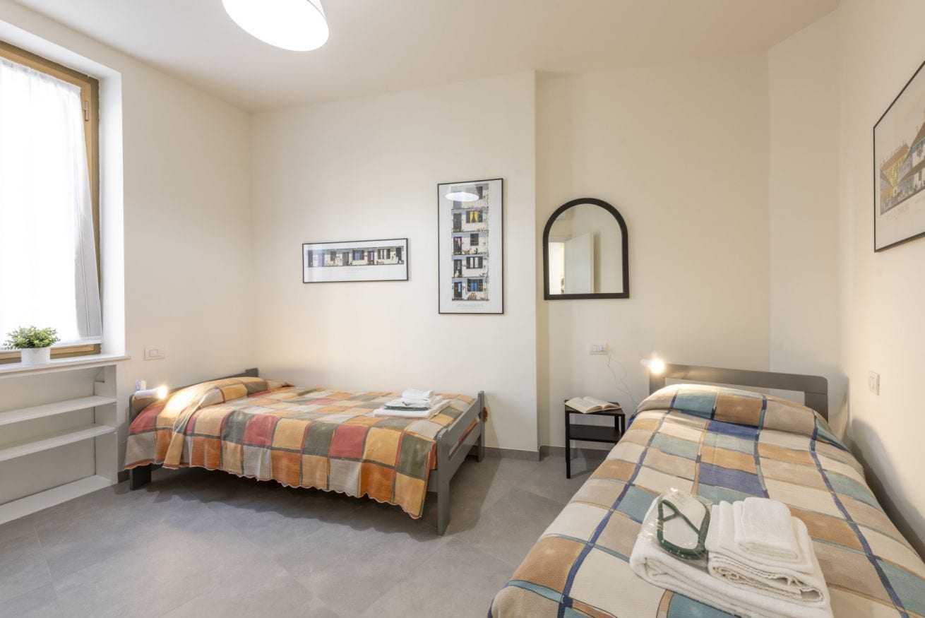 A bedroom with two beds, a window, a bedside table, and a mirror.