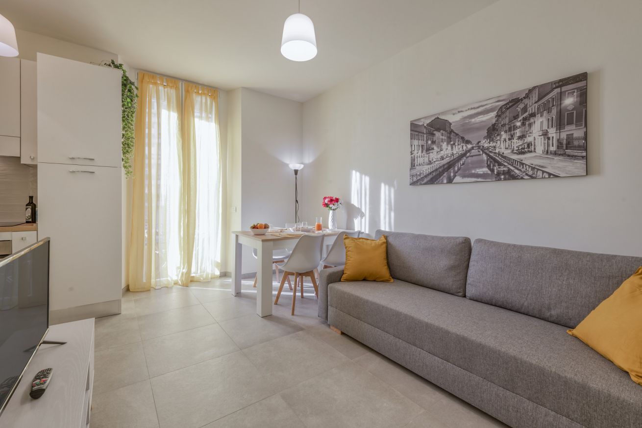 Una sala de estar moderna con un sofá gris, una mesa de comedor y una cocina con armarios blancos.