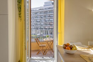 A balcony with a view of a multi-story building, a dining table with a bowl of fruit, and a chair. - BnButler - Maloja, 10 (Milano)