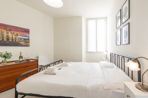 A hotel room with a bed, a wooden dresser, a nightstand, a lamp, a window, and a painting of a cityscape. - BnButler - Maloja, 10 (Milano)