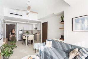 A modern living room with a grey sofa, a coffee table, and a dining area with chairs and a table. There is a kitchen in the background with white cabinets and a ceiling fan.